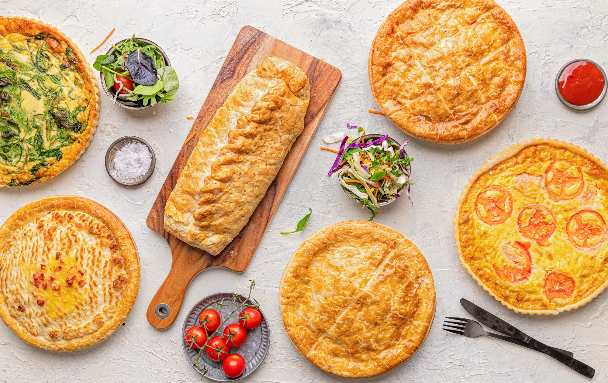 A spread of Tony's Pies products — two quiches, a sausage roll, an apple pie, a steak pie and a chicken pie laid out from above on wooden boards
